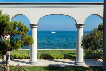 Fototapeta na wymiar backyard with arched vaults and columns with a flowerbed of green lawns and pine trees in the background is a sea horizon with a yacht into the sea, tourism background.