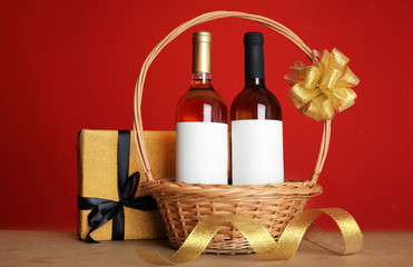 Bottles of wine in wicker basket with bow and gift on table against color background