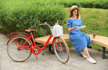 Young pretty woman reading on bench near bicycle in park