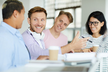 Positive enterprising young man with headphones on neck sitting at table and gesturing hand while explaining project idea to colleagues