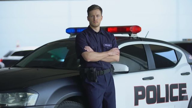 Professional policeman in uniform with crossed hands leaning on patrol car, duty