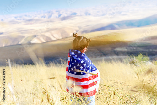 American flag. Back view little patriotic happy girl wrapping in usa national flag on beautiful landscape background. Patriotic child on wheat field. National 4 july. Memorial day.