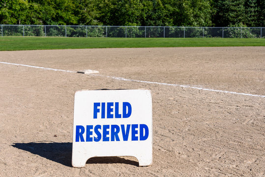 Close up of Field Reserved sign on empty local baseball field, third base and baseline, on a sunny day with woods in the background