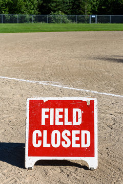 Close up of Field Closed sign on empty local baseball field, third base and baseline, on a sunny day with woods in the background