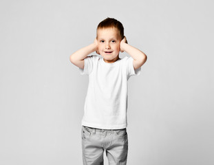 The little boy covered his ears with the palms of his hands. On a light background in a white T-shirt and gray pants
