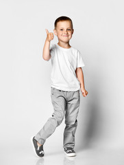 Little cute boy preschooler in a white T-shirt and gray pants on a light background, happily looking at the camera, showing a thumb sign cool