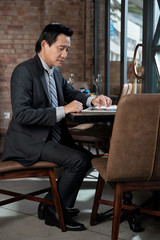 Mature Vietnamese entrepreneur in suit sitting at restaurant table and working with documents on tablet computer