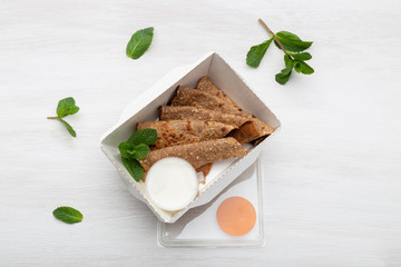 Top view of pancakes with sour cream sauce in a white lunch box lie on a white table next to the greens. Dietary snack concept.