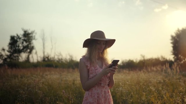 Woman In Hat Typing On Mobile At Sunset.Close Up Girl With Smartphone.Beautiful Woman Having Chat Using Smartphone Outdoors.Woman Chatting With Friends.Relaxed Girl Looking At Mobile Phone On Field.