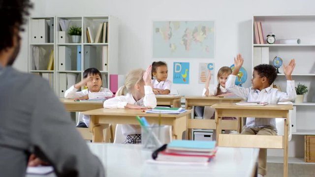 Tracking shot of unrecognizable male primary teacher sitting at his desk and asking question from cute little children smiling and raising their hands