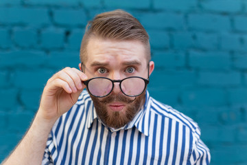 Close-up portrait of a curious surprised young man in glasses with a mustache and beard posing on a background of blue blurred brick wall. Concept of surprise and shocking information.