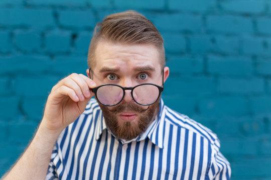 Close-up portrait of a curious surprised young man in glasses with a mustache and beard posing on a background of blue blurred brick wall. Concept of surprise and shocking information.