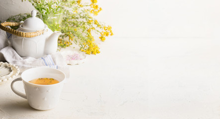 Herbal tea background with cup with yellow tea , tea pot and fresh herbs and flowers on white table...