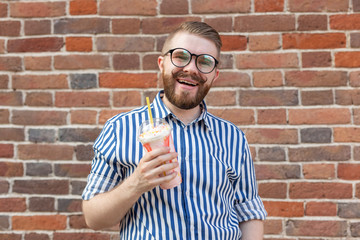 Positive young hipster guy in glasses and with a beard is holding a cocktail with a pipe posing against a brick wall. The concept of holidays and summer vacation.