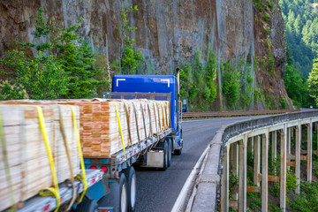 Big rig blue semi truck with two flat bed semi trailers transporting lumber running on the bridge on the road around of the mountain rock