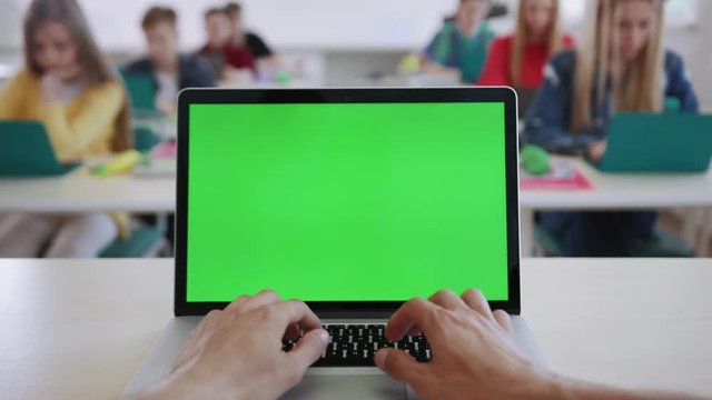Close-up of male hands of a teacher typing on laptop keyboard with mockup screen sitting in the classroom. View of clever students working on computers during a lesson in a modern school.