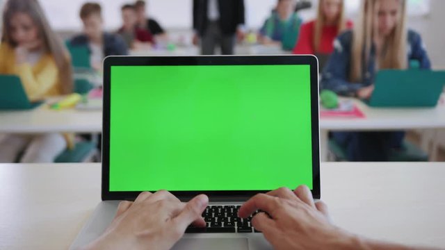 Teacher using a computer laptop with greenscreen mockup sitting in front of the class. Young students working on laptops by desks during a future technology lesson in a school.