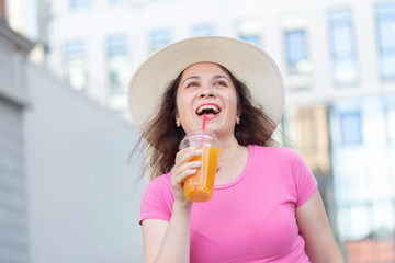 Bottom view portrait of a young beautiful cheerful woman in a hat walking around the city with juice on a warm summer day. The concept of walking through the city at the weekend.