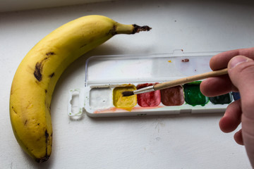 a banana and watercolor paint and brush in hand of artist on white background