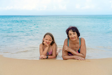 Fototapeta premium Portrait of young granddaughter and elderly grandmother look at camera posing for family picture, happy little girl and aged woman lie by the sea on a clear day.