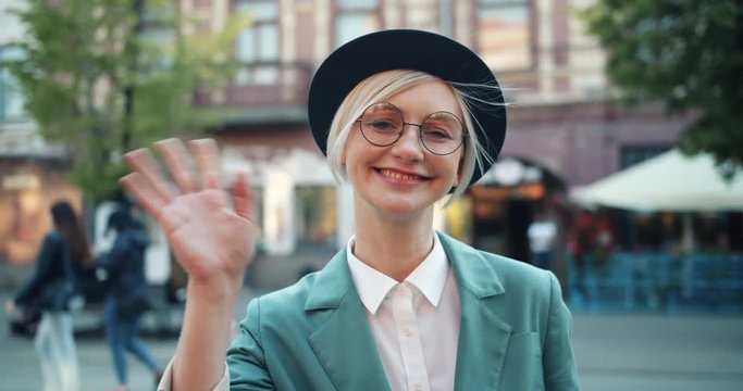Slow motion of happy girl waving hand in the street looking at camera in city standing alone greeting friends. Welcome, hello and friendly youth concept.
