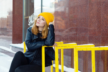 Young woman with blue eyes and blond hair in a yellow knitting hat and black leather jacket on the...