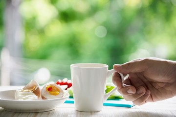 Hand hooding hot coffee cup with boiled eggs with fresh cucumber potato onion salad breakfast set with green forest background - breakfast food concept