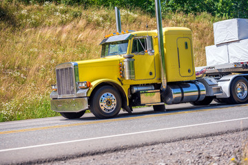 Fototapeta na wymiar Classic yellow big rig semi truck transporting cargo on flat bed semi trailer running downhill