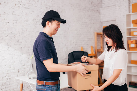 Side View Of Smiling Asian Female Signing Tablet While Taking Carton Box From Happy Delivery Man In Office