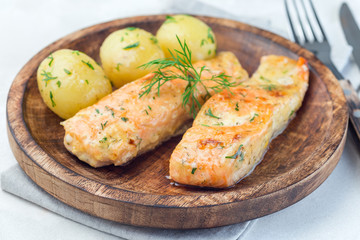 Baked salmon in creamy sauce with young boiled  potato topped with melted butter and chopped dill on wooden plate, horizontal