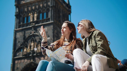 Two young women sit under the old tower and show to each other neighborhood