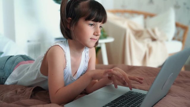 Little girl lying on bed, uses laptop, presses buttons on keyboard, slow motion