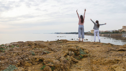 Two young women stand on stones in the sea and put their hands up