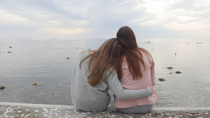 Two young women sitting on the waterfront. Huging and look away