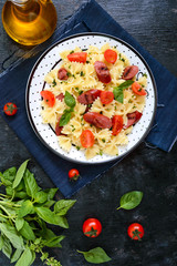 Tasty pasta farfalle with grilled sausages, fresh cherry tomatoes and basil on a plate on a black background. Top view, flat lay.