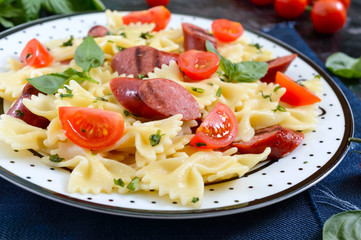 Tasty pasta farfalle with grilled sausages, fresh cherry tomatoes and basil on a plate on a black background.