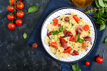 Tasty pasta farfalle with grilled sausages, fresh cherry tomatoes and basil on a plate on a black background. Top view, flat lay.