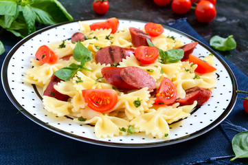 Tasty pasta farfalle with grilled sausages, fresh cherry tomatoes and basil on a plate on a black background.
