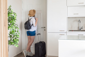 woman with suitcase and straw hat in shirt and shorts looking in peephole door waiting a taxi and ready to trip. Individual traveler.