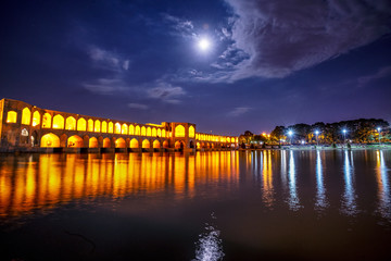 Khaju Bridge over Zayandeh river is iluminated at dusk with lights and moon in sky, Serving as a dam as well