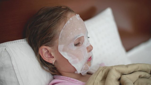 Portrait of child girl with cloth mask on the face, side view. Skin care procedure at home for teen problem skin. Moisturizing face mask fo kid.