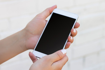 Female hand holding smartphone on white brick wall background, mockup with copy space. Phone with a blank screen for design