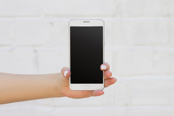 Female hand holding smartphone on white brick wall background, mockup with copy space