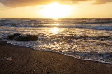 Dramatic sunrise with waves crashing over the rocky beach in El Medano, Tenerife, Canary Islands, Spain. Beautiful seascape with vibrant colors and golden haze and dynamic waves striking sandy shores.