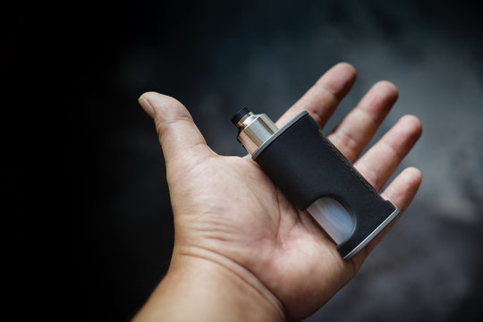 high end unregulated bottom feed squonk box mods with rebuildable dripping atomizer in hand on dark grey texture background, vaping device, vape gear, vaporizer equipment, selective focus