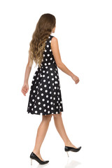 Beautiful Young Woman In Black Dotted Cocktail Dress And High Heels Is Walking. Rear Side View.