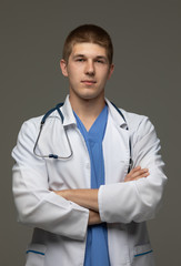 Closeup shot of young caucasian man doctor standing with crossed arms in white uniform against grey background looking perfectly confident.