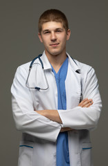 Closeup shot of young caucasian man doctor standing with crossed arms in white uniform against grey background looking perfectly confident.