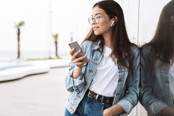 Smiling young pretty woman student wearing eyeglasses walking outdoors listening music with...