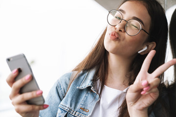 Funny young pretty woman student wearing eyeglasses walking outdoors listening music with earphones...